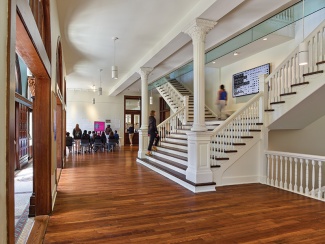 Interior of the historic Richardson Memorial Hall with polished wood floors, white columns, and a grand staircase. Students walk through the space, and a group sits in chairs at the far end.