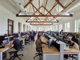 Architecture students work in a bright studio space, seated at large, long tables of computer workstations.