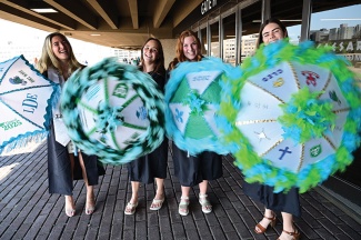 Four graduates in cap and gown outside of the Superdome twirling decorated umbrellas 