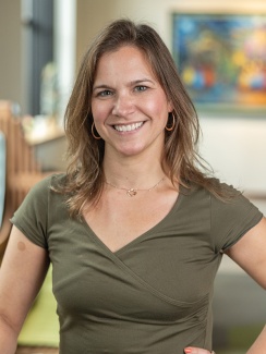 Portrait of Catherine O'Connor. Smiling woman with brown hair in a green top stands confidently indoors. Background shows abstract art and soft lighting, conveying a warm, welcoming feel.
