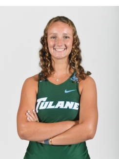 Young woman with curly hair smiling confidently, wearing a green Tulane athletic tank top, arms crossed. Studio backdrop with a focused, positive vibe.