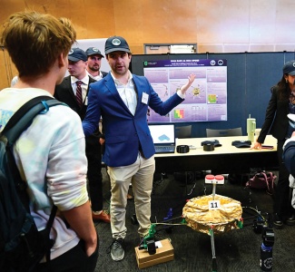 A man in a blue blazer and cap presents a space rover model at an expo. A laptop and informational poster are on the table behind him.