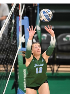Volleyball player in a green jersey, number 13, prepares to set the ball near the net, displaying focus and concentration. Indoor court setting.