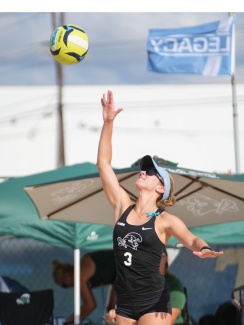 A female beach volleyball player in a black jersey jumps to hit a ball midair. She wears sunglasses and a visor. Blue sky and flags in the background.