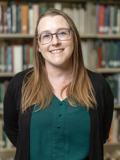 Portrait of Jacquelyne Thoni Howard. A smiling woman with glasses stands in front of a bookshelf, wearing a green blouse and black cardigan. 