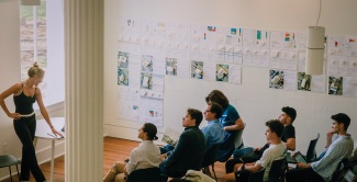 View of the first floor open space from the staircase of Richardson Memorial Hall. Seated students listen to a presentation. A gallery wall displays architectural projects.