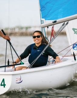 Chun Jie K. Ricci wearing sunglasses and a life jacket smiles confidently while steering a sailboat. The blue sail is raised, and the water is slightly choppy.