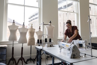 A student uses a sewing machine in a light-filled studio with large windows. Dress form dummies are in the background.