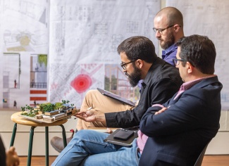 Three architecture students examine an architectural model. Architectural plans are displayed on the wall in the background.