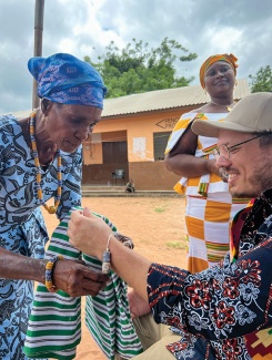 Elderly woman in blue headscarf ties a bracelet to a seated man's wrist. Woman in colorful attire smiles nearby. 
