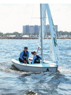 Two sailing teammates in life vests sail a small white boat on a sunny day. The water glistens, and a city skyline is visible in the background.