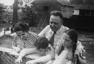 The Fenves family of four leans over a wooden fence on their farm. The mother, father, young son, and daughter  look into an enclosure. Behind them are geese, a fenced yard, and rustic farm buildings with brick walls and wooden roofs.