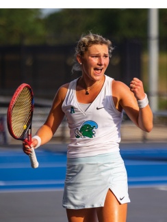 A female tennis player wearing a white outfit passionately celebrates with a fist pump on an outdoor court, holding a red racket.