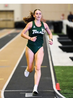 Athlete in green uniform runs on an indoor track, displaying focus and determination. Her hair flows behind, with a blurred background of a sports facility.