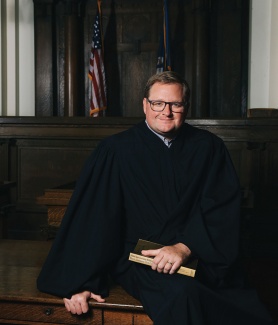 Justice Cade Cole in black judicial robes sits in a formal courtroom setting, wearing glasses and holding a book.
