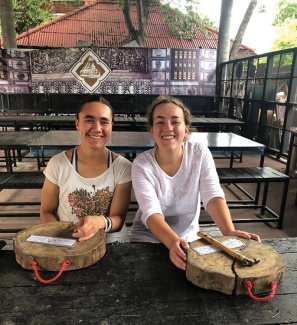 Two smiling students sit side by side at a wooden table, each holding a drum. Background features a mural and trees, conveying a relaxed, cheerful atmosphere.