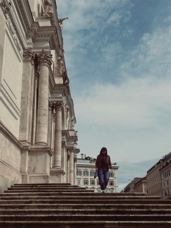 A person walks down a grand stone staircase beside a historic building with ornate columns and sculptures. The sky is partly cloudy, adding depth to the scene.