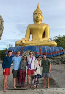 A group of six people stands smiling in front of a large golden Buddha statue. The setting is outdoors on a clear day, conveying a joyful and serene mood.