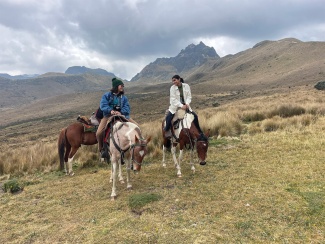 Two people ride horses on a grassy mountain landscape under a cloudy sky. They seem relaxed, engaged in conversation, with rugged peaks in the background.