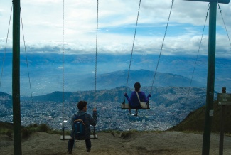 Two people on swings overlook a vast cityscape and mountains under a cloudy sky. The scene conveys a sense of freedom and adventure.