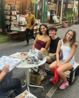 Three people sit smiling at an outdoor market, posing for an artist sketching them. Stalls display books in the background, creating a lively atmosphere.