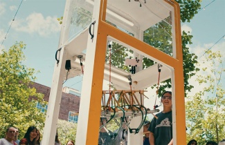 A smiling person wearing safety goggles stands near a large outdoor testing machine with yellow wooden framing, under a clear sky with green trees.