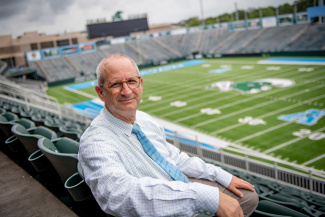 A man in glasses and a light checked shirt sits in a stadium's empty stands, overlooking a green football field. The sky is overcast, adding a calm tone.