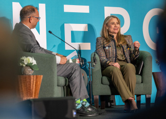 A woman in a patterned blazer speaks on a panel beside a man in a suit. They're seated on green chairs with a turquoise backdrop, conveying a professional and engaging atmosphere.
