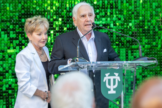 A man in a black suit speaks at a podium with a woman in white beside him. The background is a shimmering green with a Tulane logo. The mood is formal.