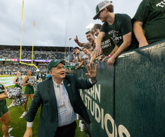 A man in a suit and cap high-fives enthusiastic fans at a football game. Cheerleaders stand nearby. The atmosphere is lively and celebratory.
