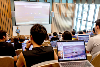 A lecture hall filled with students using laptops. A speaker stands at the front near a projected presentation. The atmosphere is focused and engaged.