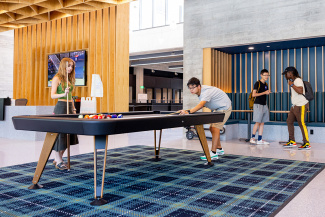 Two people focus intently playing pool in a modern, open lobby with wooden accents. The atmosphere is relaxed, while two others chat in the background.
