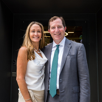 A smiling woman in a white dress and a man in a gray suit with a teal tie stand closely together, posing happily in front of a doorway.