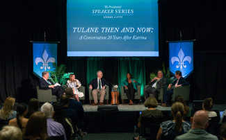 Panel discussion with six speakers seated on stage, under a screen displaying "Tulane Then and Now: A Conversation 20 Years After Katrina." Audience attentively watches.