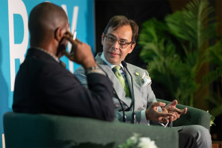 Two men seated and engaged in a discussion at a formal event. Indoor setting with plants.