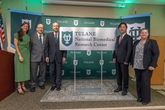 A group of five people stands smiling in front of a green backdrop featuring the Tulane logo. A sign reads "Tulane National Biomedical Research Center." The scene conveys professionalism and celebration.
