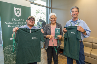 Three people stand smiling, holding green Tulane T-shirts. A Tulane National Biomedical Research Center banner is behind them, conveying a celebratory mood.