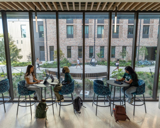 Students study at high tables by large windows, with a view of a campus building and walking students outside. The atmosphere is focused and studious.