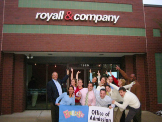 A group of smiling people stands outside a brick building with a sign reading “royall & company.” They hold a Tulane University Office of Admission banner.
