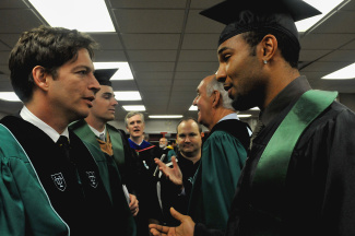 Graduation scene with Harry Connick Jr. and Matt Forte in caps and gowns engaging in animated conversation. The atmosphere is lively and celebratory, highlighting achievement.