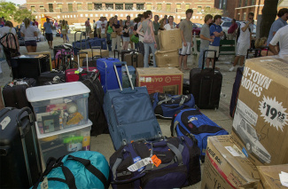 A bustling scene of college move-in day, featuring cardboard boxes and suitcases piled high. Students and families gather in the background, engaging in conversations.