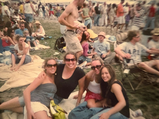 Four women smiling and sitting on a blanket at an outdoor event, surrounded by a crowd enjoying a sunny, festival-like atmosphere.