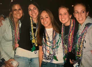 Five young women smiling, wearing colorful bead necklaces and casual attire. They appear joyful, suggesting a festive, celebratory atmosphere.