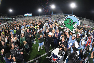 Fans celebrate on a football field at night, confetti falling, person holding a Tulane Angry Wave logo.