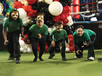 Four people in green shirts, three with tiger masks, crouch on a green floor.