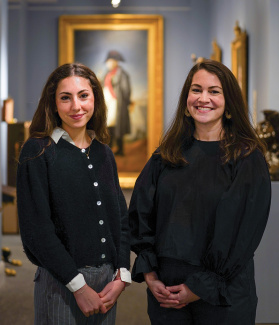 Two women smiling in an art gallery, a classical portrait painting behind them.