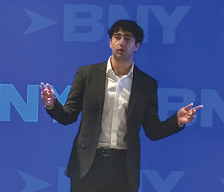 A person in a dark suit gestures while speaking on stage against a blue backdrop with blurred 'BNY' logos. The scene conveys professionalism and engagement.
