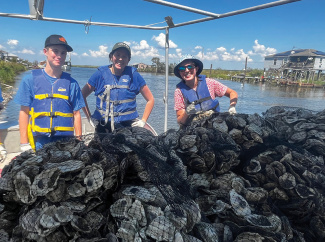 Three people in life vests on a boat loaded with oysters, a sunny bay behind.
