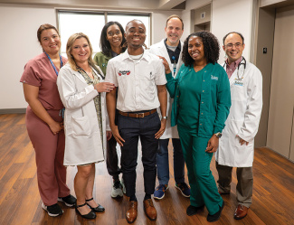 Eight smiling healthcare professionals in scrubs and lab coats in a hospital hallway.