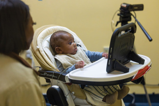 Baby in beige high chair looks right, woman observes. Camera on tripod in background.